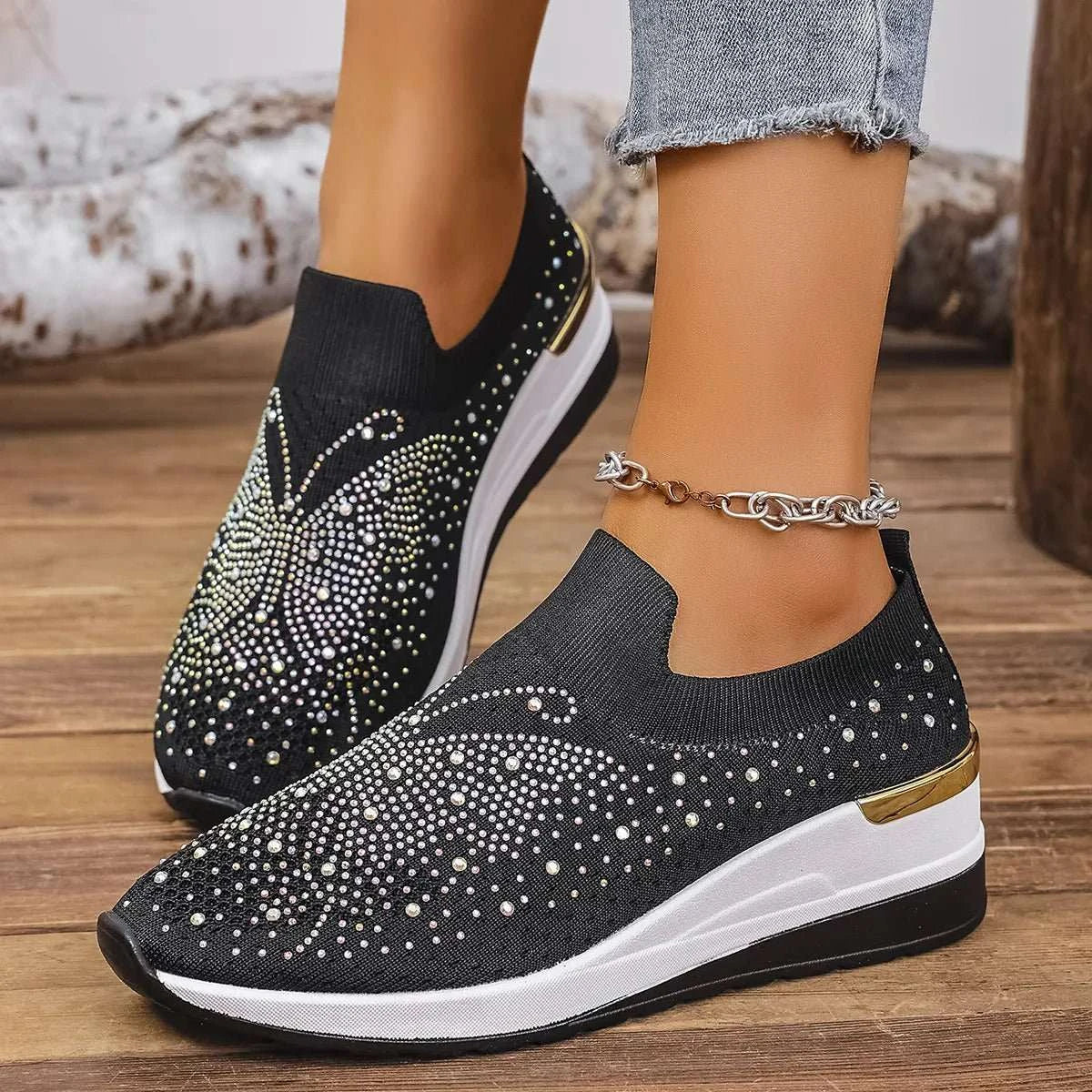 Black rhinestone wedge sneakers with butterfly design, mesh material, mid heels, and white sole worn with ankle bracelet and frayed denim jeans