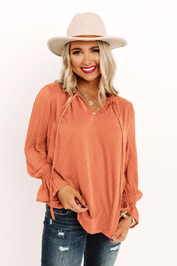 Woman wearing rust-colored Swiss dot tie neck blouse with flounce long sleeves and casual chic style paired with distressed jeans and beige hat