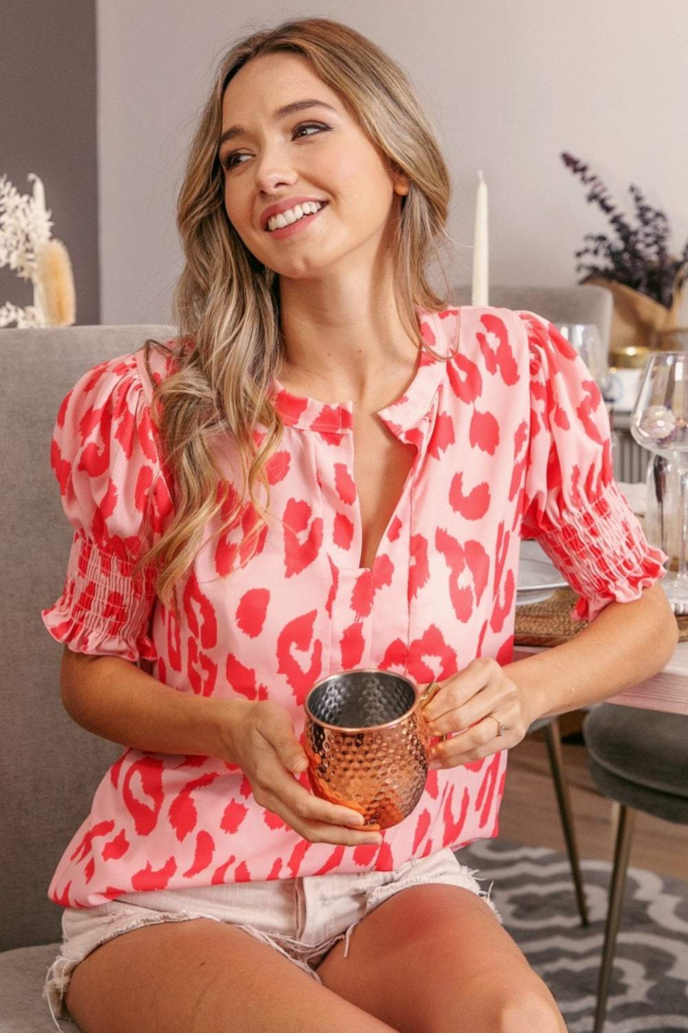 Woman wearing a pink and red BiBi Leopard Smocked Short Sleeve Blouse holding a copper mug sitting indoors