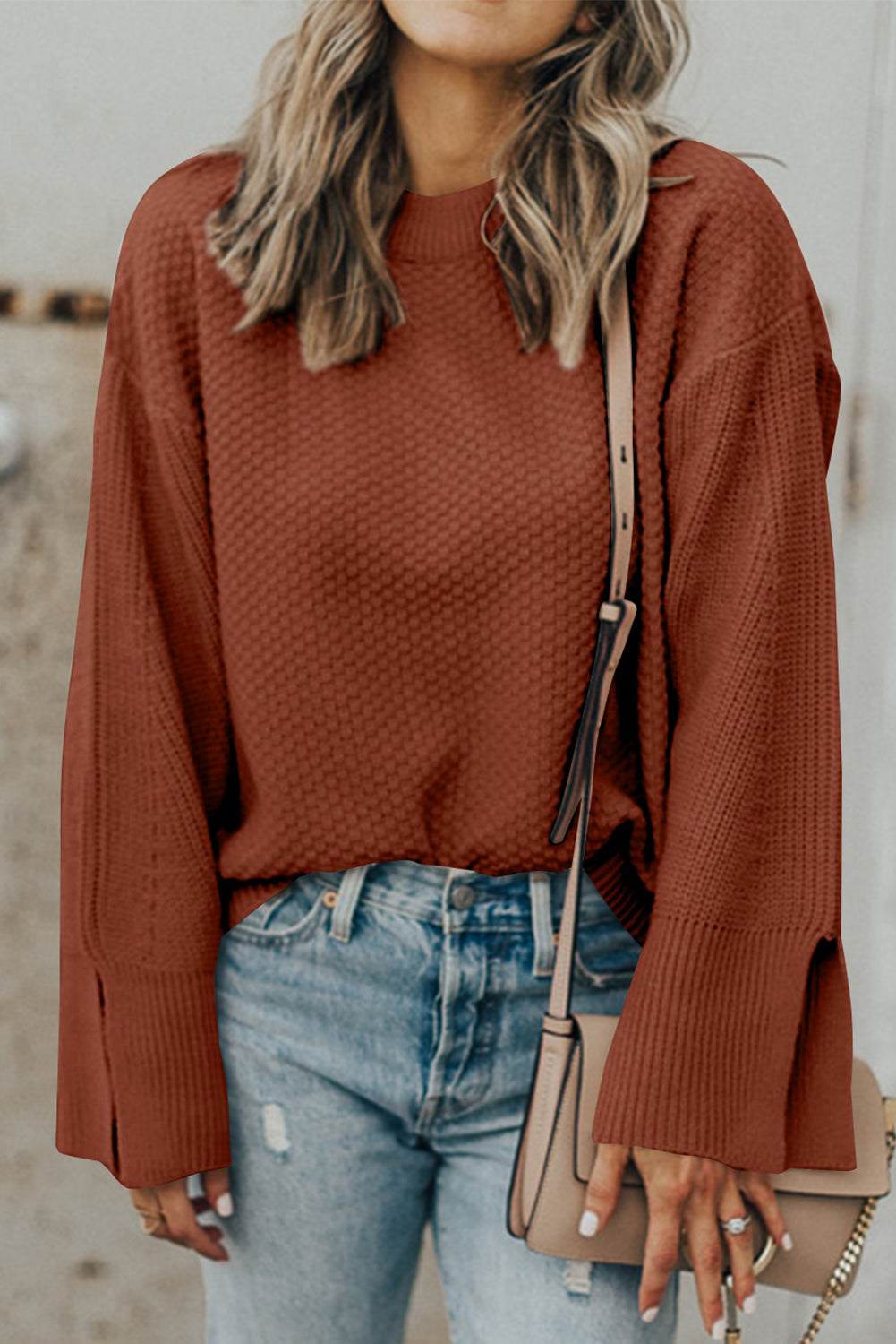Textured round neck long sleeve sweater with slits, in a warm brown shade, paired with blue jeans and a beige handbag.