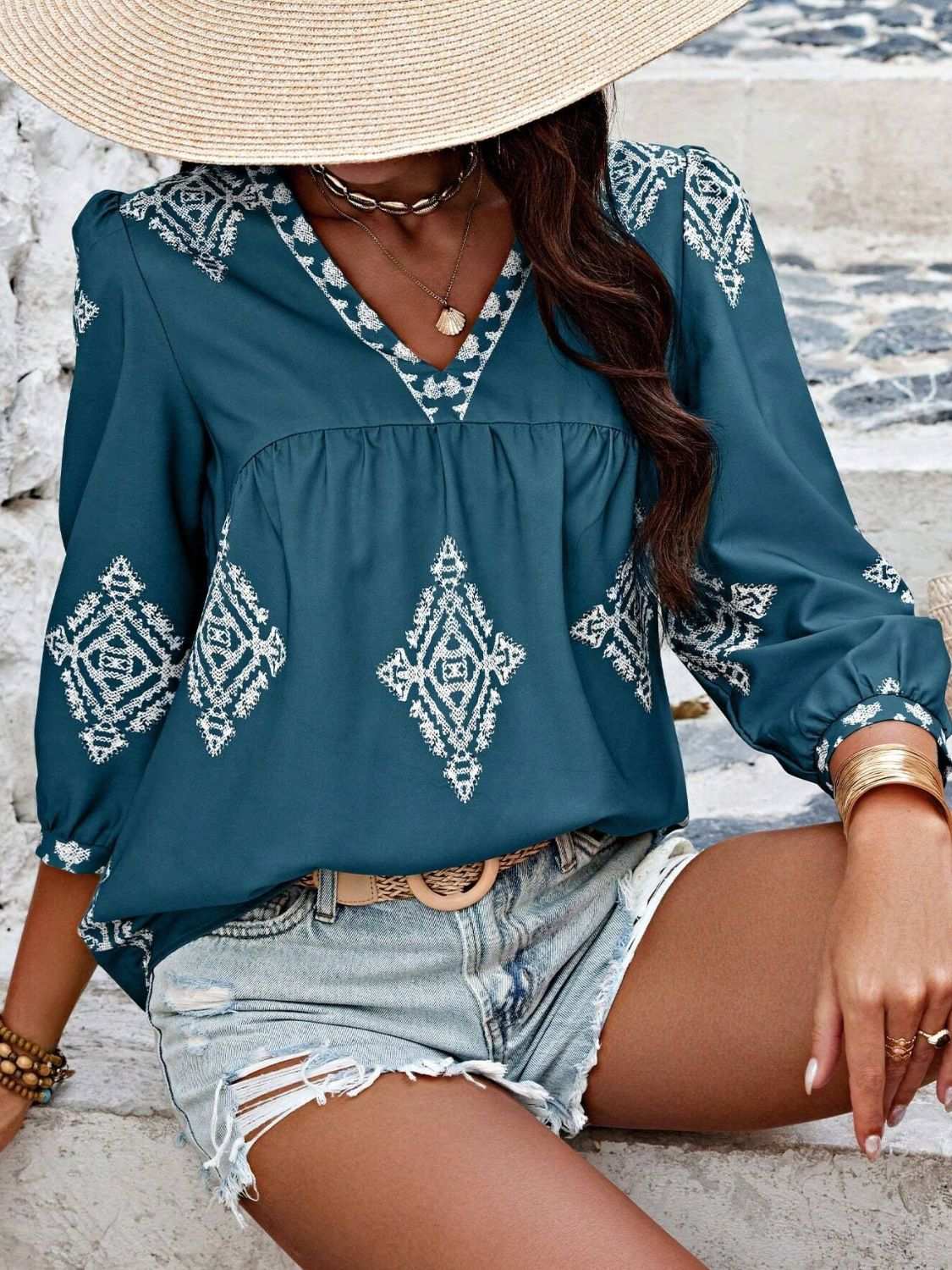 Blue printed V-neck three-quarter sleeve blouse with white geometric patterns, styled with denim shorts and a wide-brimmed hat.