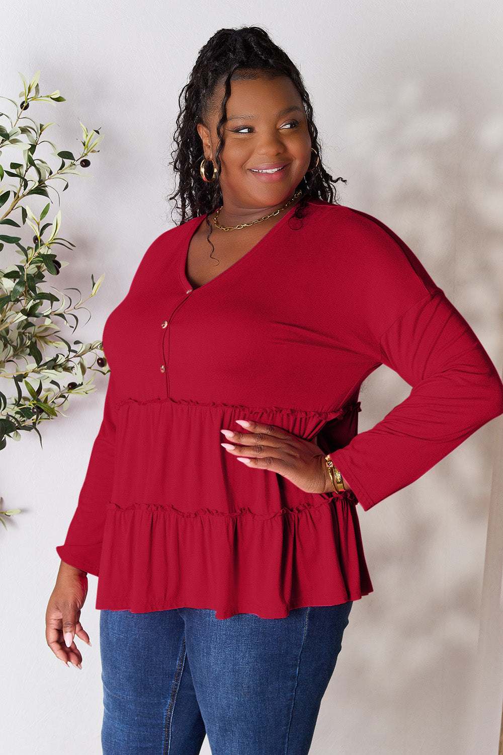 Red half button long sleeve ruffle hem blouse on a smiling woman standing next to a plant.