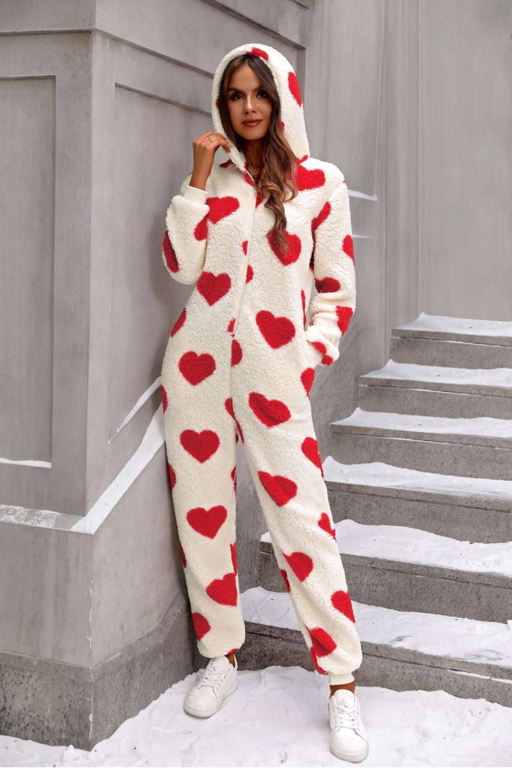 Woman wearing fuzzy heart zip up hooded lounge jumpsuit with red hearts on white fabric, casual relaxed fit polyester onesie