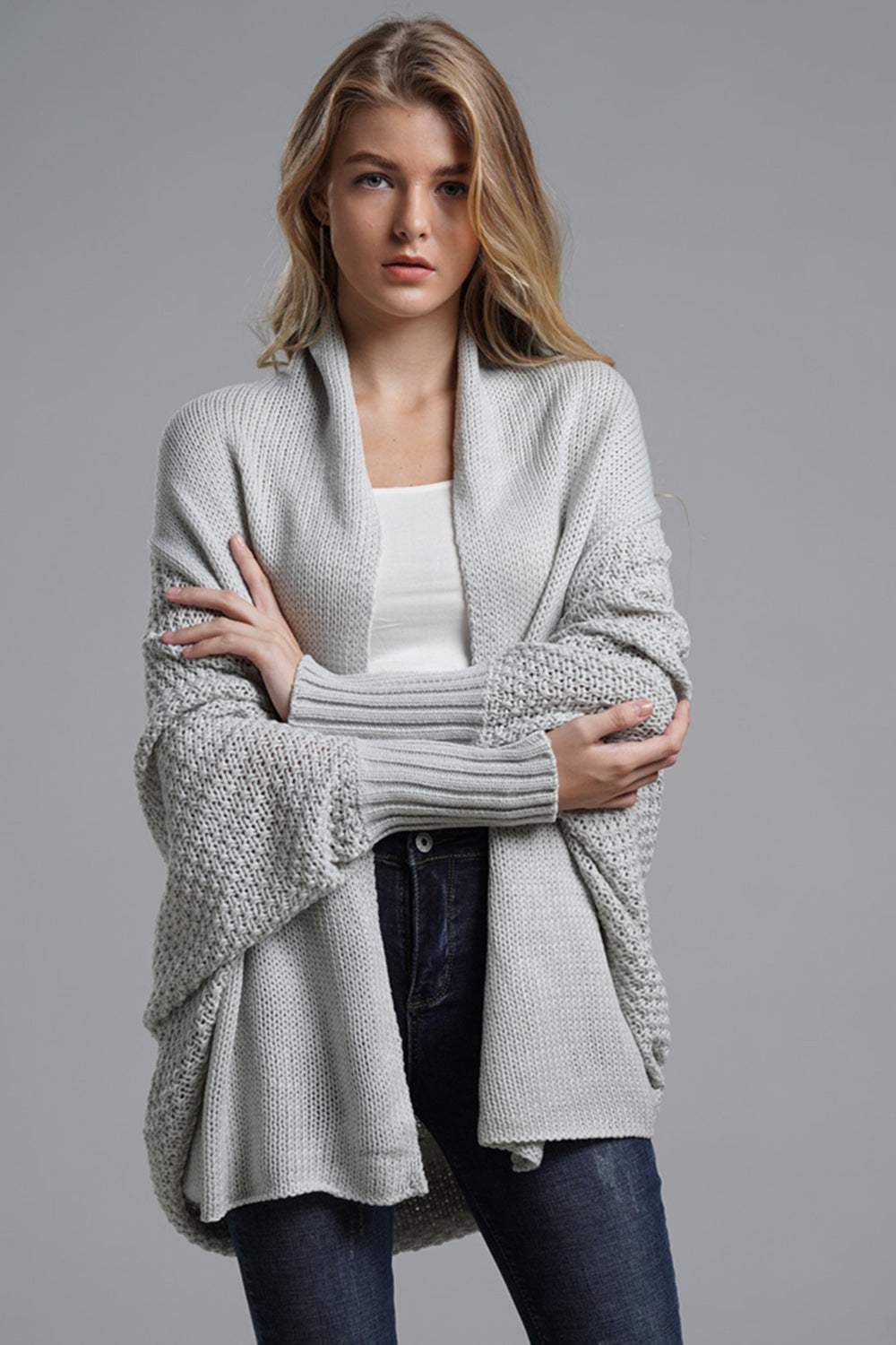Woman wearing a light gray open front batwing sleeve cardigan with textured knit and ribbed cuffs over a white top and jeans