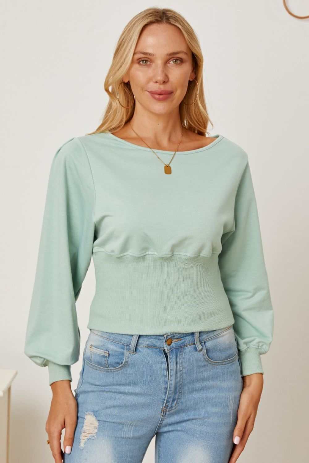 Woman wearing a light green boat neck lantern sleeve blouse with long puff sleeves and fitted waist paired with blue jeans.