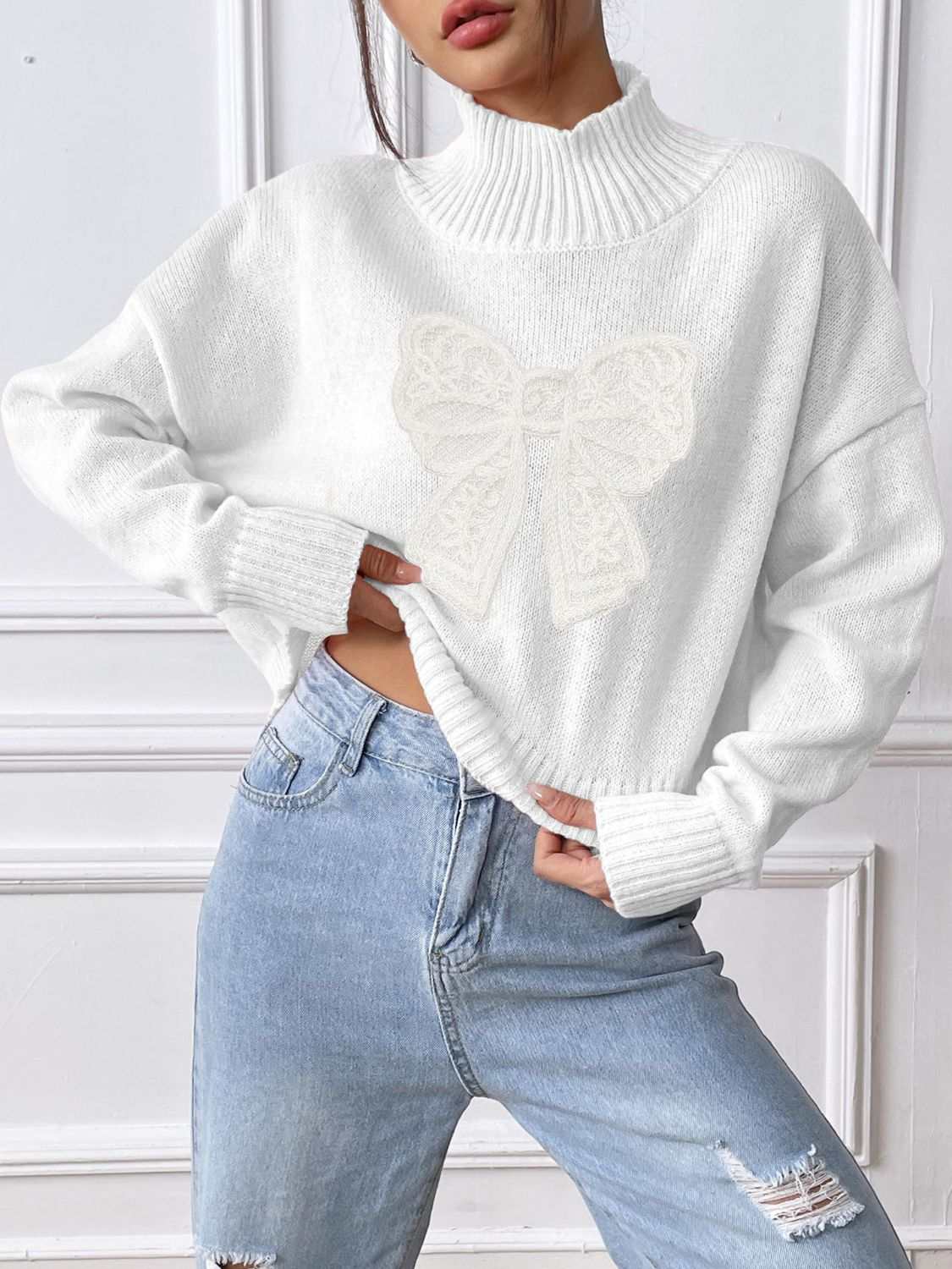 White bow turtleneck long sleeve sweater made from 100% polyester, slightly stretchy, paired with blue jeans.