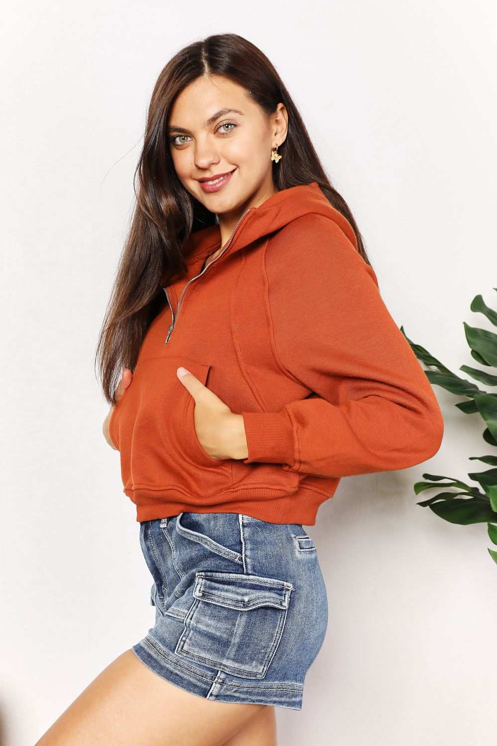Woman wearing Double Take Half-Zip Long Sleeve Hoodie in rust color with denim shorts.