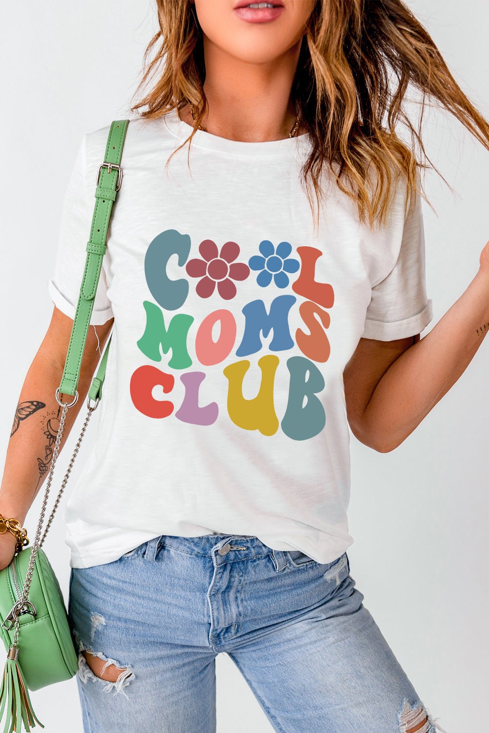 COOL MOMS CLUB round neck short sleeve t-shirt featuring colorful lettering, paired with jeans and a green bag.