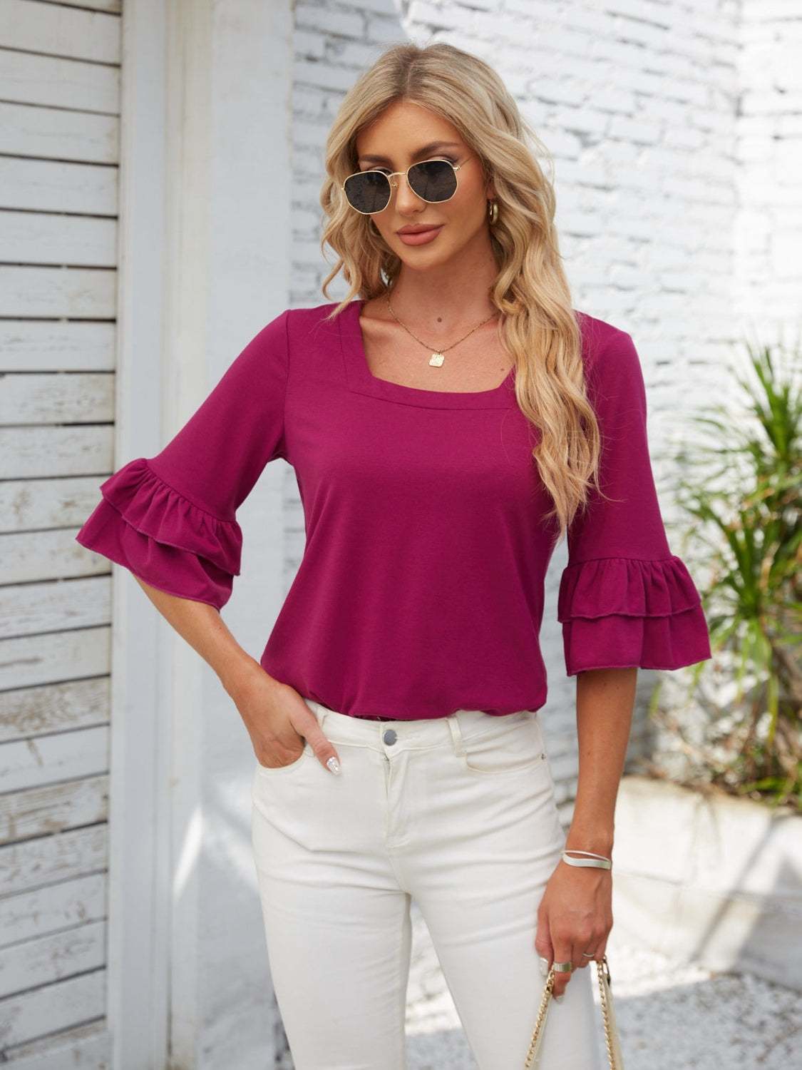 Woman wearing a magenta ruffled square neck half sleeve blouse with tiered sleeves and white pants outdoors