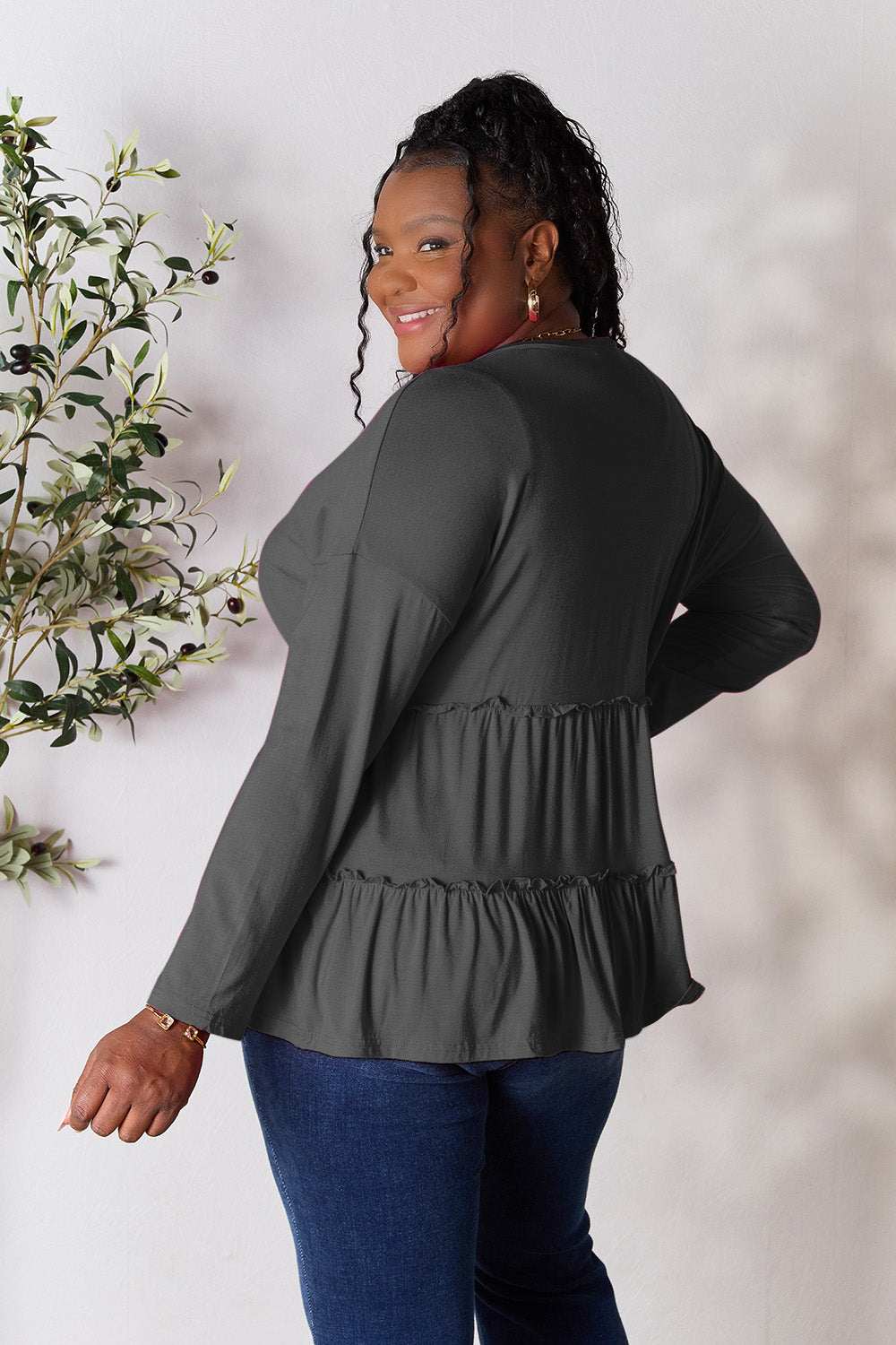 Half button long sleeve ruffle hem blouse in dark color, worn by a smiling woman facing sideways, with plant decor in the background.