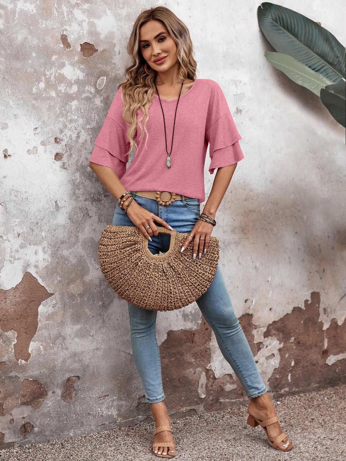 Woman wearing a pink V-neck half sleeve blouse with jeans and holding a woven bag.