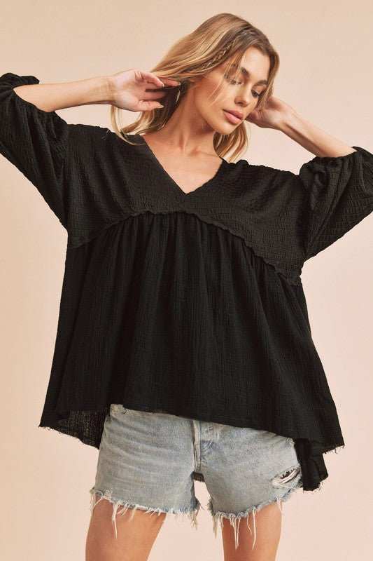 Woman wearing black Aemi + Co high-low frayed hem V-neck blouse with ruched sleeves and distressed denim shorts