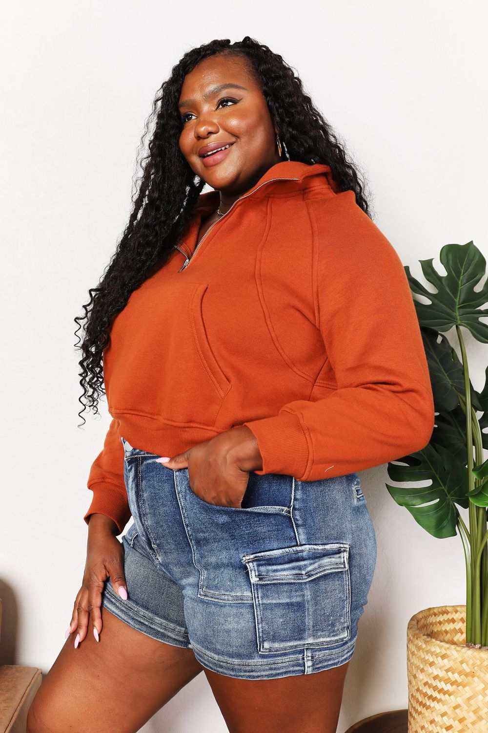 Plus-size woman wearing Double Take Half-Zip Long Sleeve Hoodie in solid color with raglan sleeves and collared neckline, paired with denim shorts.