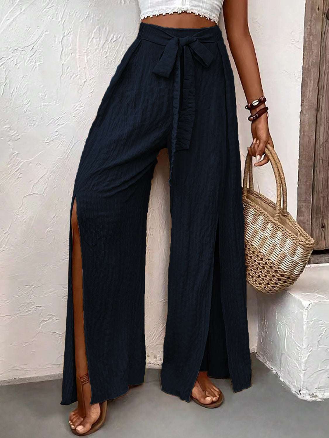 Wide leg pants with slits, honey-tied design, made from polyester and elastane, worn with sandals and a woven bag.
