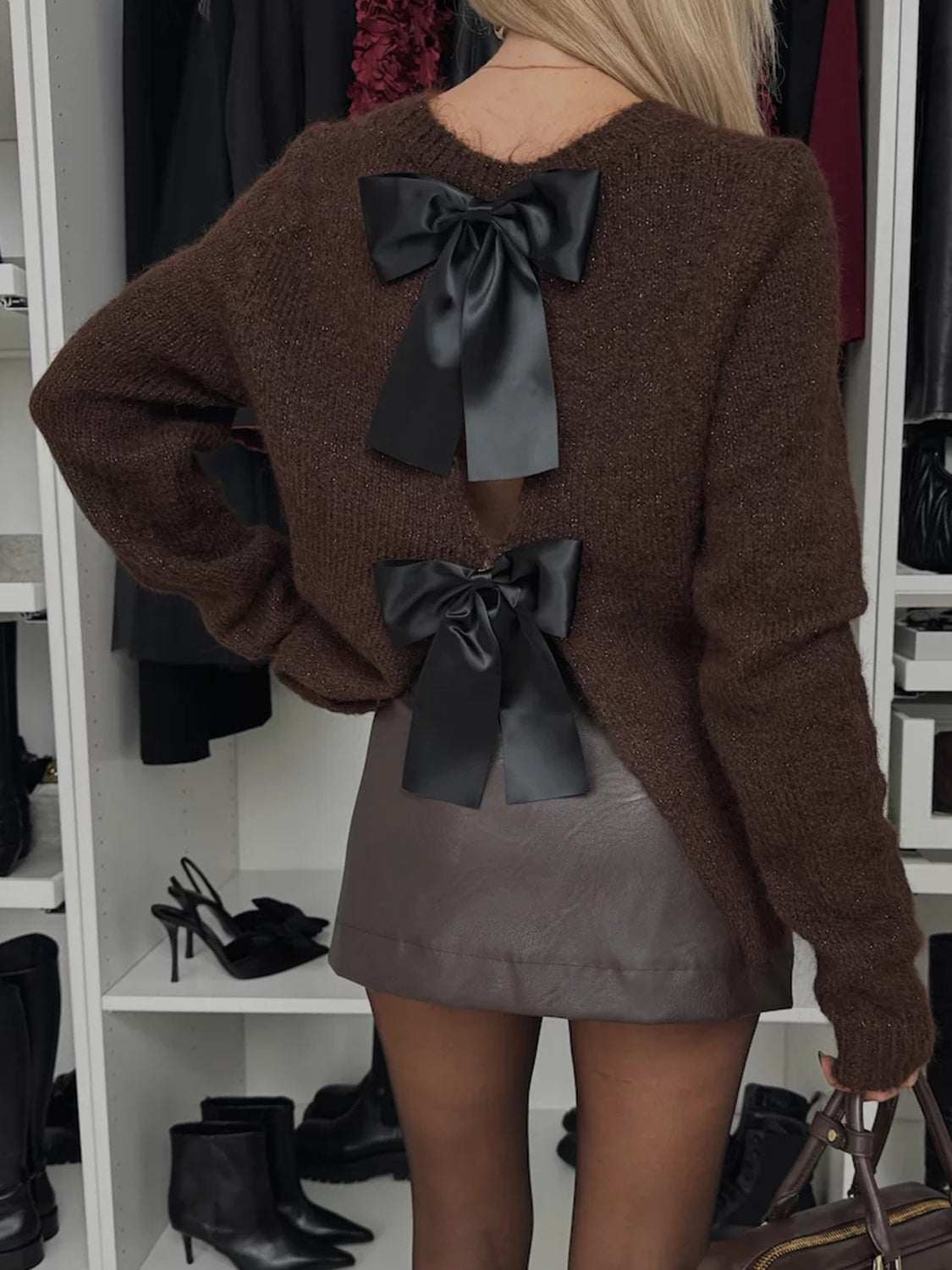 Bow slit back round neck sweater in brown with black ribbon accents, paired with a leather skirt in a closet setting.