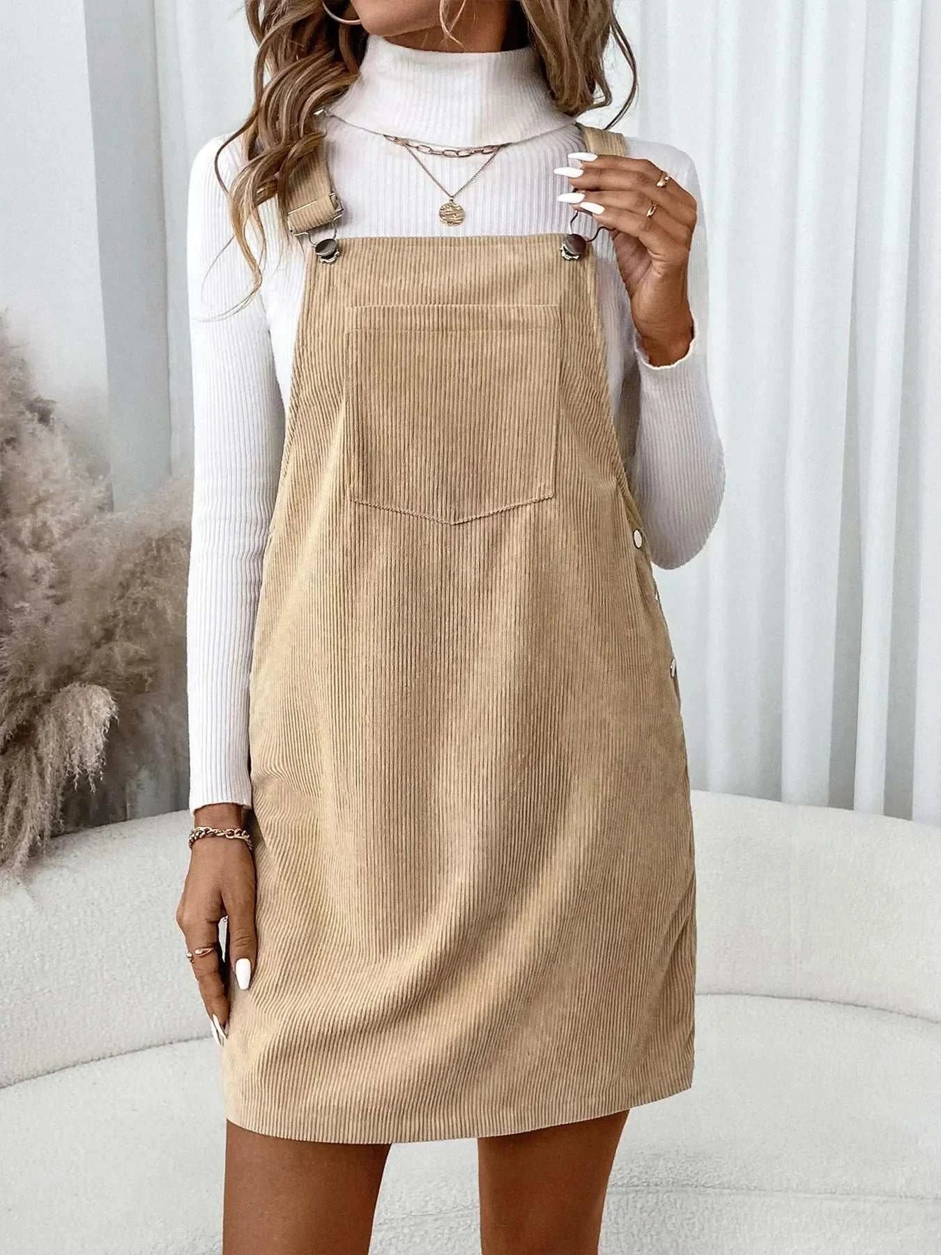 Beige corduroy overall dress with front pocket and button details, worn over a white turtleneck sweater.