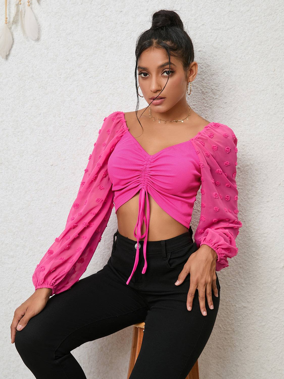 Woman wearing pink Swiss dot drawstring cropped blouse with long puff sleeves and black pants against a white textured background