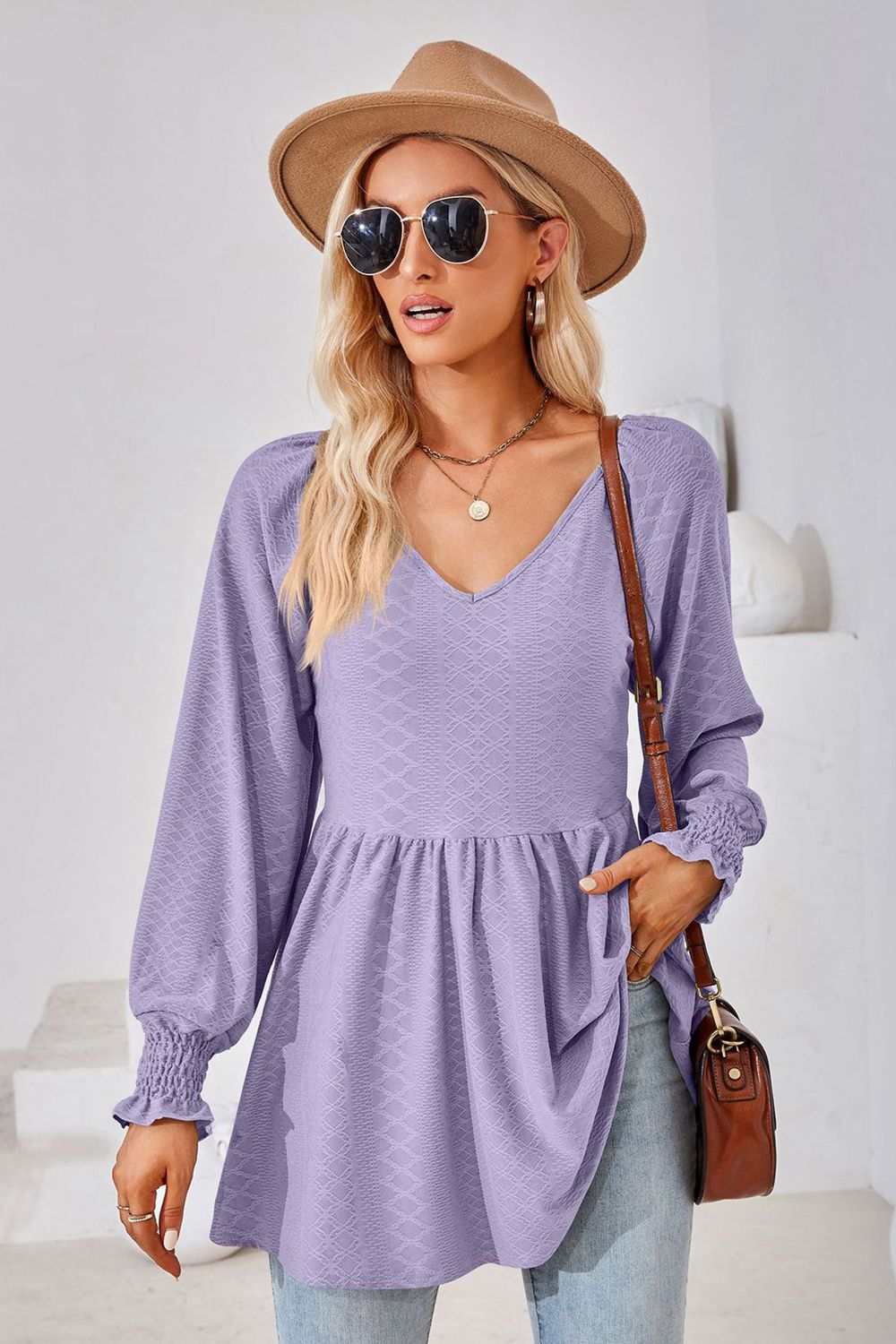 Woman wearing lavender V-neck lantern sleeve blouse with long puffed sleeves and smocked cuffs, paired with sunglasses, hat, and casual jeans.